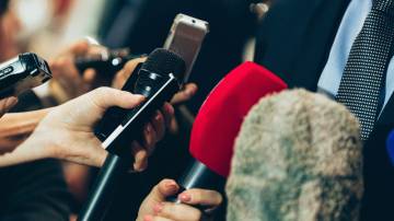 Vários jornalistas entrevistando homem de terno. Na imagem é possível ver diversos microfones apontados para o homem.