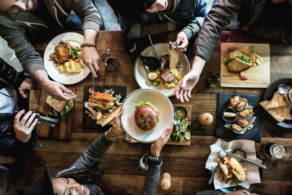 Mesa de restaurante com diversos pratos e pessoas sentadas. Uma mulher passa um prato com um hambúrguer para o outro lado da mesa