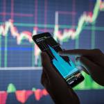 Mão segura um celular e consulta um gráfico em frente a um painel de movimentação de ações em Bolsa - mercado fracionário