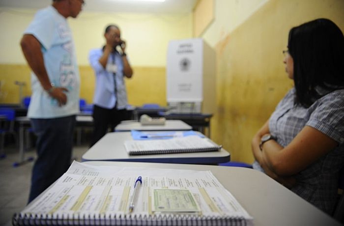 Mesários reunidos em local de votação, perto de urna eletrônica, durante eleições no Brasil (Foto: Agência Brasil)